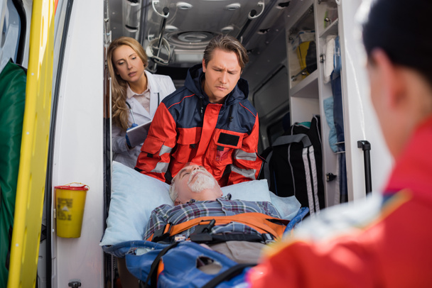 Enfoque selectivo de los paramédicos que sostienen al paciente en la camilla cerca del médico con una tableta digital en un automóvil ambulancia  - Foto, imagen