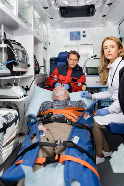 Выборочный фокус врача, смотрящего в камеру рядом со старшим пациентом и парамедиком в машине скорой помощи  - Фото, изображение