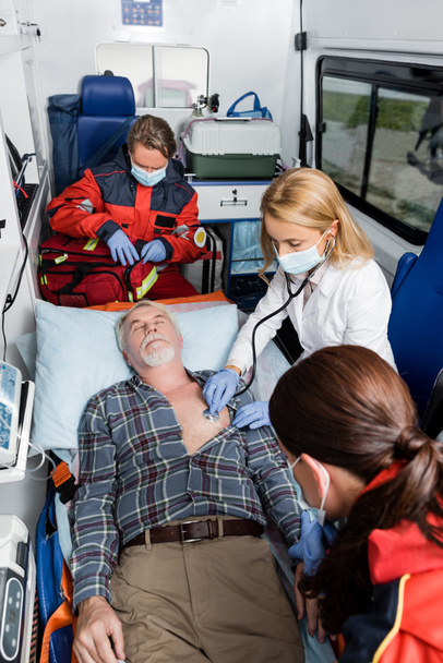 Selective focus of doctor in medical mask examining patient with stethoscope near paramedics in ambulance car  - Photo, Image