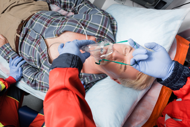 Selectieve focus van paramedicus in latex handschoenen met zuurstofmasker op zieke patiënt in ambulance auto  - Foto, afbeelding