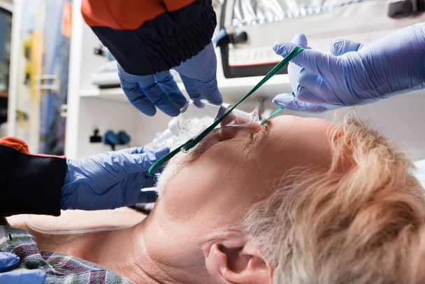 Επιλεκτική εστίαση του παραϊατρικού προσωπικού σε γάντια λατέξ φορώντας μάσκα οξυγόνου στον ασθενή σε ασθενοφόρο auto  - Φωτογραφία, εικόνα