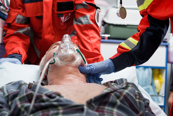 Selective focus of paramedic in latex glove checking pulse of senior patient in oxygen mask near colleague during first aid  - Valokuva, kuva