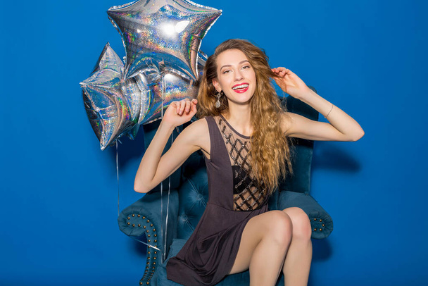 excited young hipster beautiful woman, evening dress, trend fashion style, blue background, sitting in chair, silver balloons, smiling, happy, celebrating birthday - Photo, image