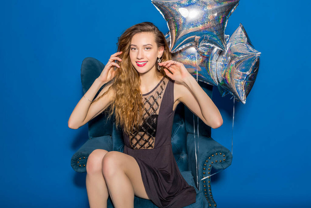 pretty young hipster beautiful woman, evening dress, trend fashion style, blue background, sitting in chair, silver balloons, smiling, happy, celebrating birthday - 写真・画像
