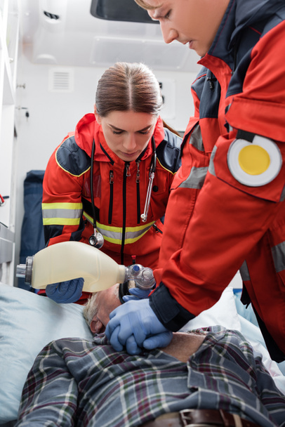 Enfoque selectivo de paramédico concentrado haciendo reanimación cardiopulmonar a paciente mayor cercano a colega en coche ambulancia  - Foto, Imagen