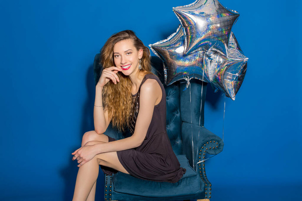 cool young hipster beautiful woman, evening dress, trend fashion style, blue background, sitting in chair, silver balloons, smiling, happy, celebrating birthday - Foto, imagen
