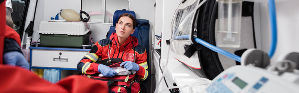 Panoramic shot of paramedic with first aid kit sitting near colleague in ambulance car   - Фото, зображення