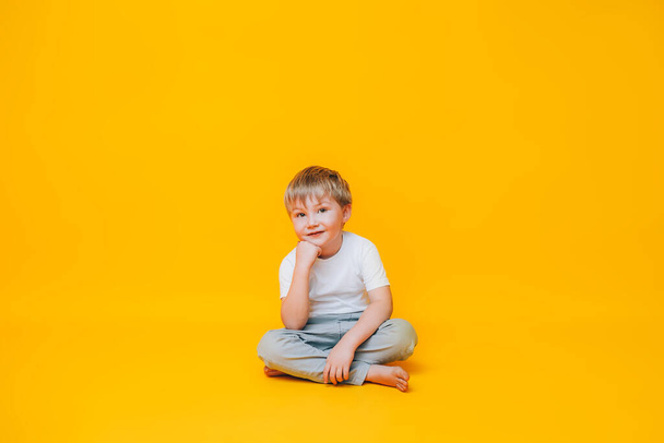 Töprengő kisfiú fehér pólóban ül a sárga háttér. - Fotó, kép