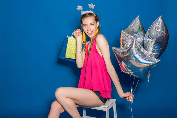 cute young hipster beautiful woman, colorful outfit, trend fashion style, blue background, holding shopping paper bags, silver star balloons, presents, smiling, happy, celebrating birthday - Foto, afbeelding