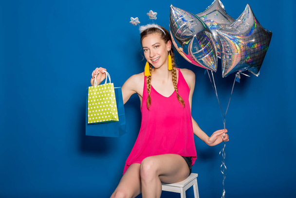 cute young hipster beautiful woman, colorful outfit, trend fashion style, blue background, holding shopping paper bags, silver star balloons, presents, smiling, happy, celebrating birthday - Photo, image