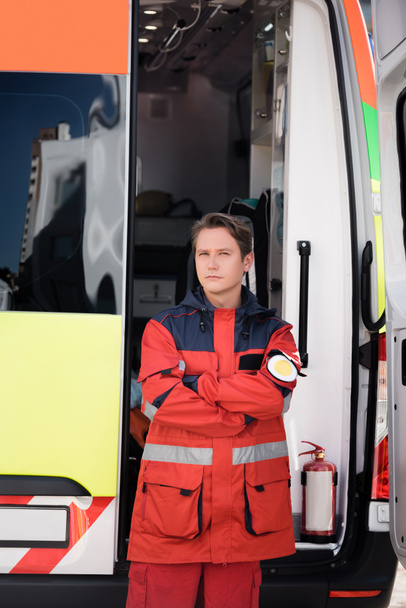 Paramédico com braços cruzados olhando para a câmera perto de carro de ambulância ao ar livre  - Foto, Imagem