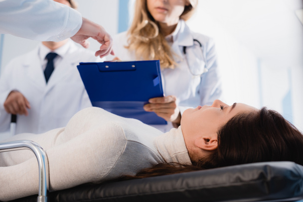 Селективный фокус пациента с закрытыми глазами, лежащего рядом с доктором, указывающим на планшет коллеге в клинике  - Фото, изображение
