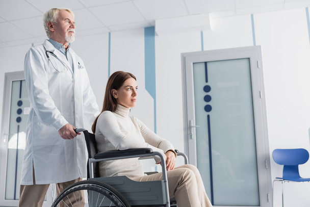 Selective focus of woman sitting in wheelchair near senior doctor in white coat in clinic  - Фото, изображение