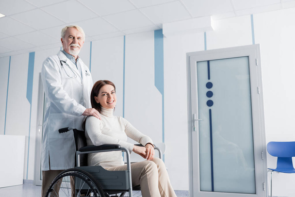 Patient in wheelchair looking at camera near elderly doctor in clinic  - Foto, imagen