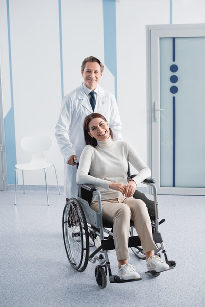 Doctor looking at camera while standing near patient on wheelchair in clinic  - Foto, Bild