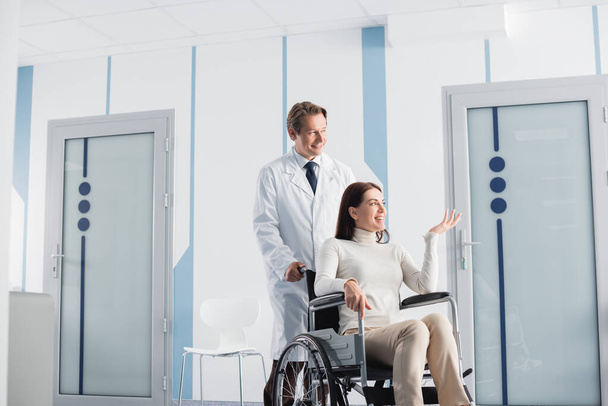 Brunette woman in wheelchair pointing with hand while sitting in wheelchair near doctor in hospital  - Photo, Image