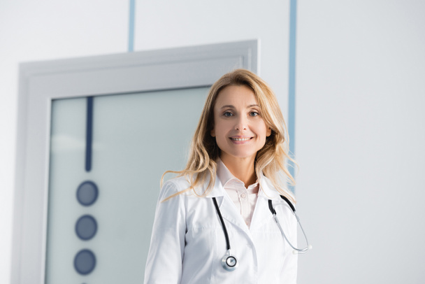 Blonde Ärztin im weißen Mantel blickt im Krankenhaus in die Kamera  - Foto, Bild