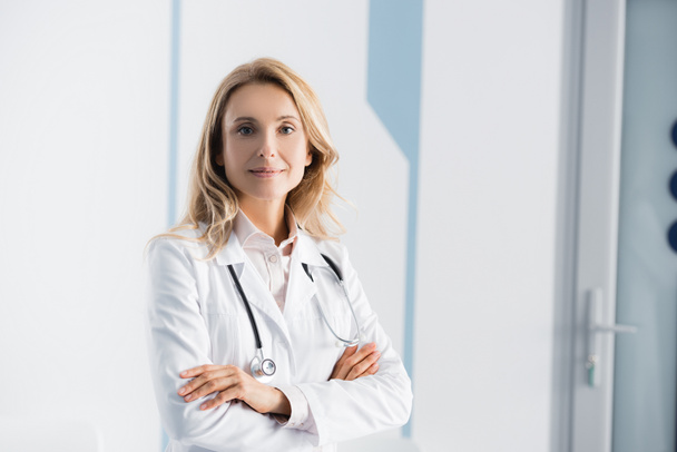 Blond arts met stethoscoop kijken naar camera in kliniek  - Foto, afbeelding