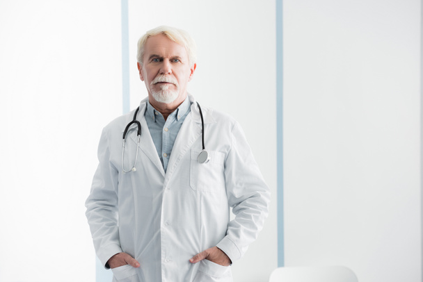 Starszy lekarz z rękami w kieszeniach patrzący przed kamerą w szpitalu  - Zdjęcie, obraz