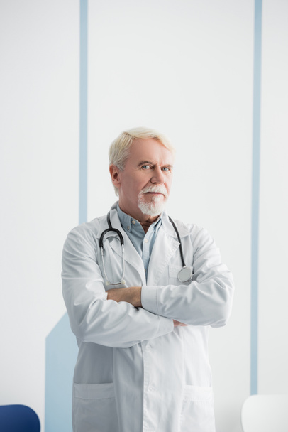 Starší lékař se zkříženýma rukama při pohledu do kamery v nemocnici  - Fotografie, Obrázek