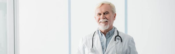 Foto panorámica del médico de edad avanzada con estetoscopio mirando a la cámara en la clínica  - Foto, Imagen