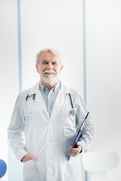 Starszy lekarz ze schowkiem patrzący na kamerę w szpitalu  - Zdjęcie, obraz