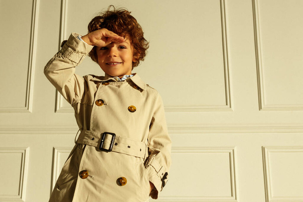 Stylish portrait of a pretty happy curly kid posing frontal in rain coat, looking down, isolated on a white background. - Φωτογραφία, εικόνα