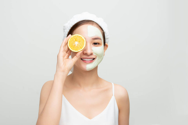Beautiful woman is getting facial clay mask and holding orange pieces on white background. Beauty, body care and spa concept - Photo, image