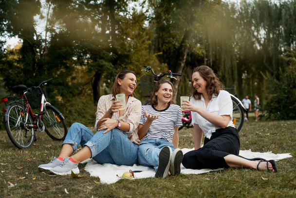 Three beautiful happy young women close friends relax in green park on a blanket. Have fun laughing and chatting on the green grass. - Fotoğraf, Görsel