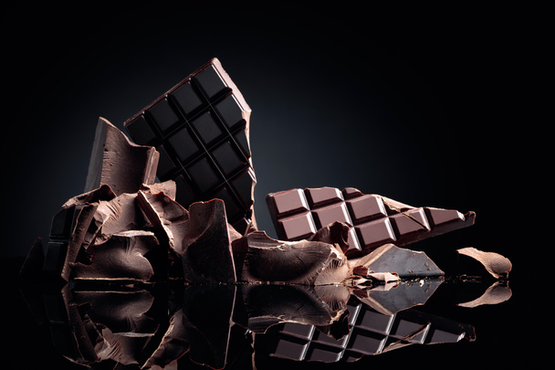 A broken chocolate bar and pieces of dark chocolate on a black reflective background. Copy space. - Photo, Image