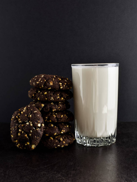 A stack of chocolate chip cookies with a nut and a full glass of milk on an old vintage table, dark background. Delicious snack, home cooking, close-up. - Foto, imagen