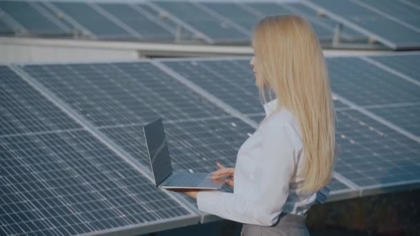 Meisje met laptop bij blauwe zonnepanelen. Vrouw in wit formeel shirt gebruiken laptop op zonnecellen achtergrond. Zon hernieuwbare energie. Groene elektriciteit in uw huis - Video