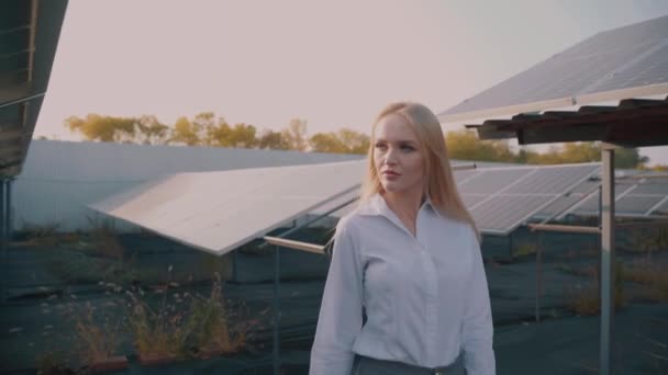 Nainen kävelee lähellä aurinkopaneelit rivi maassa auringonlaskun ja katso aurinkokennoja. Naissijoittaja käyttää virallista valkoista paitaa. Ilmainen sähkö kotiin. Planeetan kestävyys. Vihreä energia. - Materiaali, video