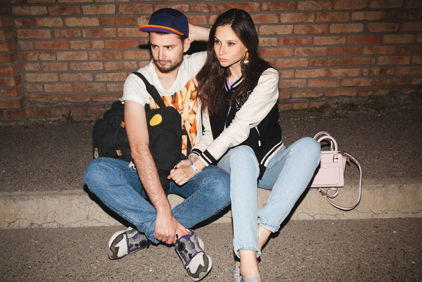 young stylish hipster couple in love, swag outfit, jeans, embrace, cool accessories, sitting on ground against brick wall, happy, having fun - Foto, Bild
