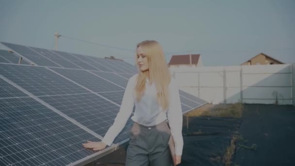 Nainen kävelee lähellä aurinkopaneelit rivi maassa auringonlaskun ja kosketa aurinkokennoja. Naissijoittaja käyttää virallista valkoista paitaa. Ilmainen sähkö kotiin. Planeetan kestävyys. Vihreä energia. - Materiaali, video