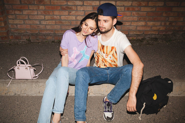 young stylish hipster couple in love, swag outfit, jeans, embrace, cool accessories, sitting on ground against brick wall, happy, having fun - Фото, изображение