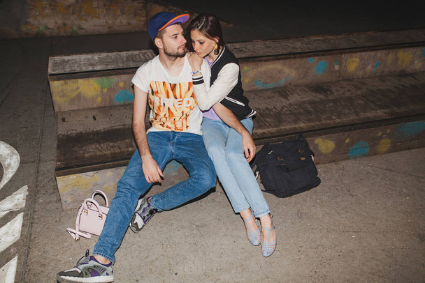 крутая молодая стильная хипстерская пара влюбленная, наряд, джинсы, объятия, крутые аксессуары, сидящая счастливая, веселая - Фото, изображение
