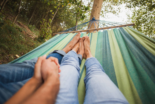 man and woman in love lying in hammock embracing, holding hands together, legs, close-up details, romance, autumn vacation, forest, relaxing, barefoot, jeans, denim outfit - Foto, imagen