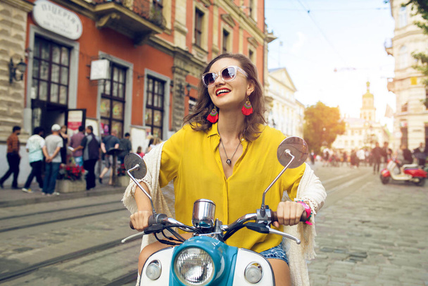 young beautiful hipster woman riding on motorbike city street, summer europe vacation, traveling, smiling, happy, having fun, sunglasses, stylish outfit, adventures, positive, waving hand - Photo, image