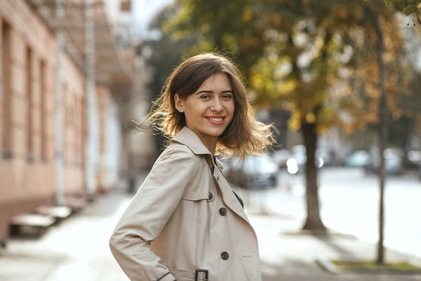 Fashion straatportret van een vrolijke brunette vrouw met natuurlijke make-up draagt elegante beige jas, lopen door de herfst stad. Ruimte voor tekst - Foto, afbeelding