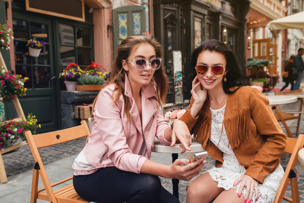 two young beautiful hipster women sitting at cafe, stylish trendy outfit, europe vacation, street style, happy, having fun, smiling, sunglasses, looking at smartphone - Photo, Image