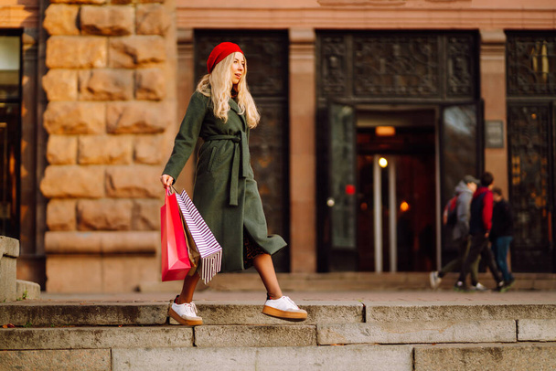 Portret młodej eleganckiej kobiety noszącej stylowy czerwony beret trzymającej torby na zakupy na ulicy miasta. Kobieta mody lubi zakupy. Konsumpcjonizm, zakupy, sprzedaż, Koncepcja Black Friday. - Zdjęcie, obraz