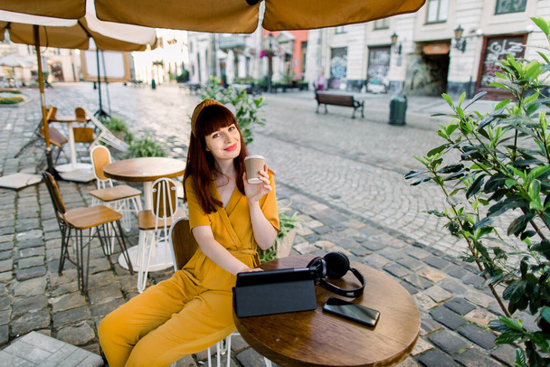 Top angle portrait of attractive young Caucasian red haired woman drinking takeaway coffee, sitting at the table at outdoor city cafe, smiling to camera, while working on tablet - Photo, Image