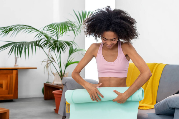 Mujer afro rodando estera de yoga het después de practicar en casa - Foto, Imagen