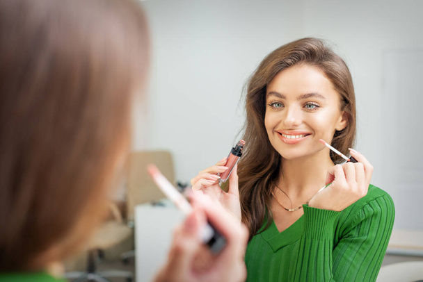 Belle jeune femme souriante caucasienne appliquant du brillant sur les lèvres en regardant dans le miroir - Photo, image
