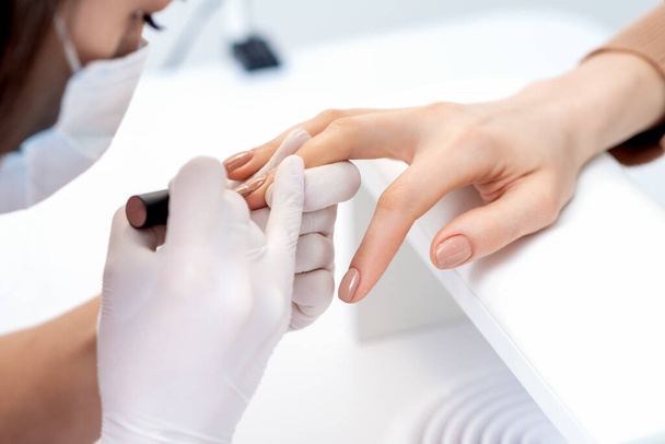 Μανικιούρ master σε προστατευτικά γάντια εφαρμόζοντας μπεζ βερνίκι νυχιών σε γυναικεία νύχια στο σαλόνι ομορφιάς - Φωτογραφία, εικόνα