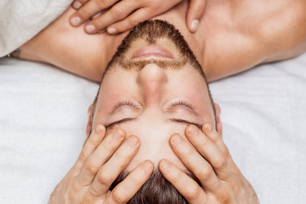 Bonito jovem recebendo massagem na cabeça relaxante por quatro mãos de dois massagistas no centro de spa de saúde - Foto, Imagem