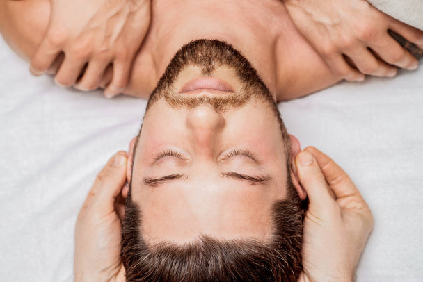 Bonito jovem recebendo massagem na cabeça relaxante de dois massagistas em quatro mãos em um centro de spa de saúde - Foto, Imagem