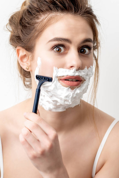 Beautiful young caucasian woman shaving her face by razor on white background. Pretty woman with shaving foam on her face - Foto, Imagem