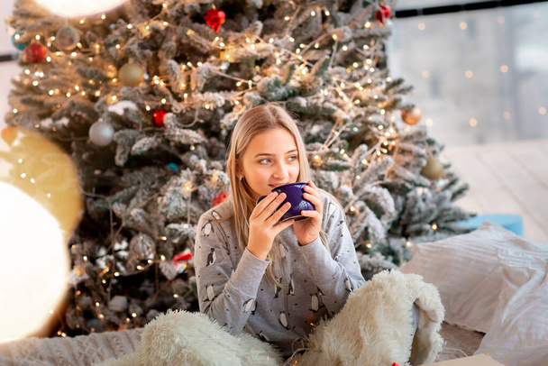 Dreamful cute teen girl near Christmas tree with cup of cacao. Kid wearing pajama dreaming near tree in the morning. Merry Christmas and Happy Holidays concept - Fotoğraf, Görsel